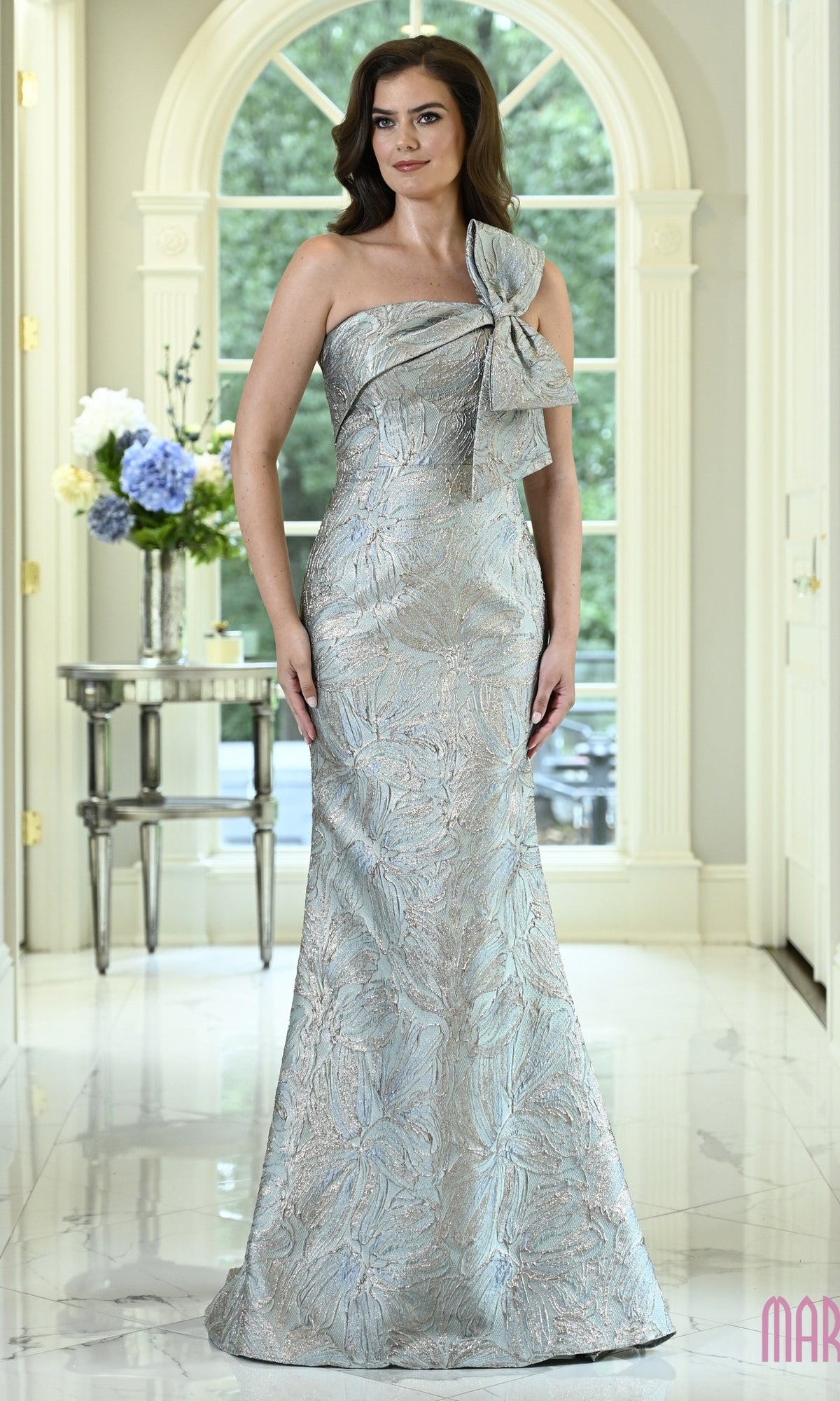 A woman wears the Marsoni MV1429, a sage-colored, floor-length floral jacquard one-shoulder evening gown with a large bow detail.